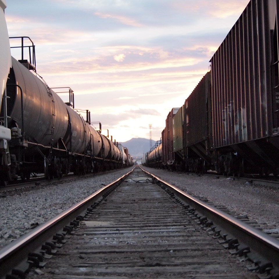 Railroad tracks between two rows of freight train cars at sunset, with mountains in the background.